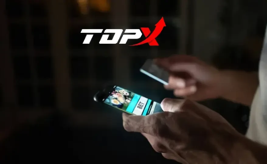 Duas mãos segurando um celular com um jogo na tela, sobre fundo escuro com o logotipo da TopX